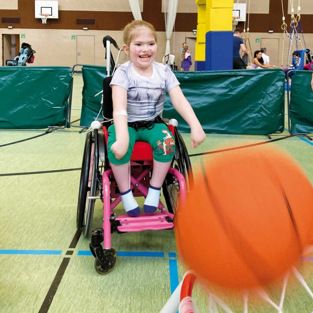 Ein Mädchen im Rollstuhl wirft lachend einen Basketball.