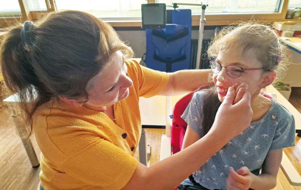 Eine Frau hilft einem Mädchen im Rollstuhl ihr Gesicht einzucremen.